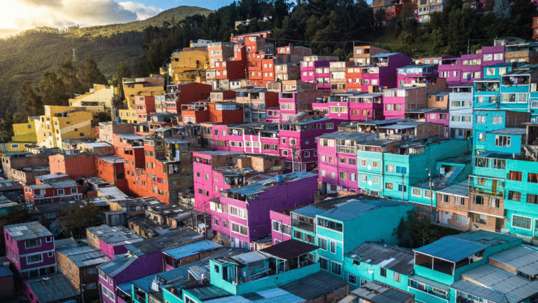 A vibrant hillside neighborhood in South America with colorful houses painted in bright shades of pink, turquoise, red, and yellow, surrounded by lush green hills under a partly cloudy sky