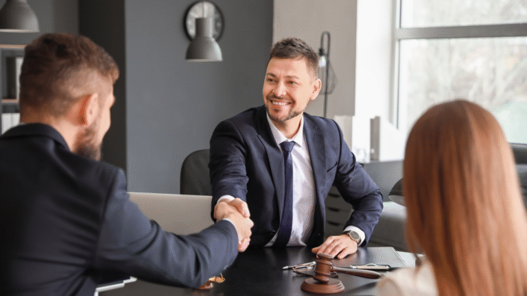 Smiling male lawyer in a suit shaking hands with a male client during a legal consultation, with a female client seated nearby and a gavel on the desk.