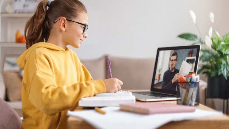 Young student attending an online tutoring session at home, taking notes while learning from a virtual tutor on a laptop.