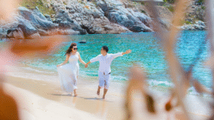 Romantic couple enjoying their honeymoon walk on a tropical beach with turquoise water and scenic rocky cliffs in the background.
