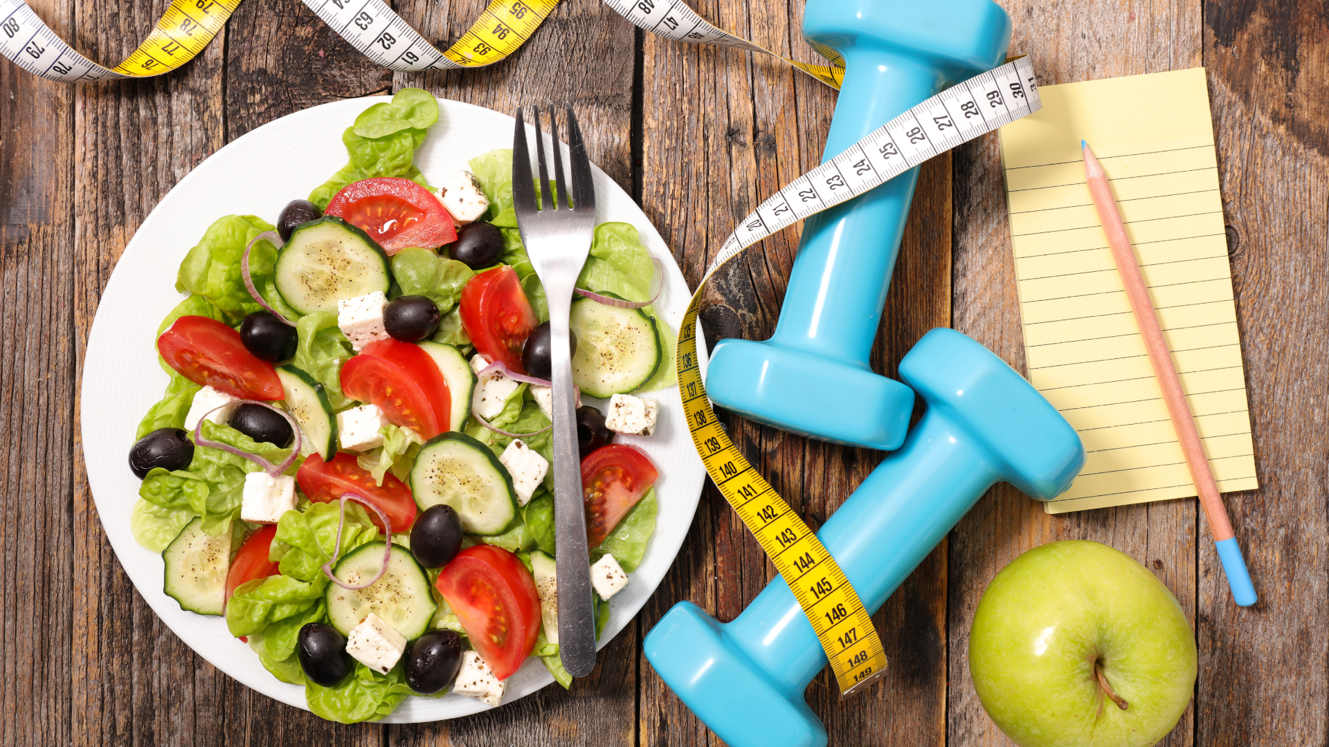 Healthy post workout food including a fresh vegetable salad with tomatoes, cucumbers, feta cheese, and olives, surrounded by blue dumbbells, a green apple, measuring tape, and a notepad for fitness tracking.