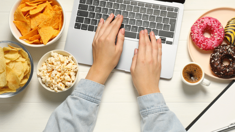 Health habits at work: person typing on a laptop surrounded by chips, popcorn, donuts, and coffee, illustrating poor snacking choices.