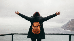 Traveler enjoying winter travel with arms wide open, wearing a black coat and backpack near a misty coastal mountain view