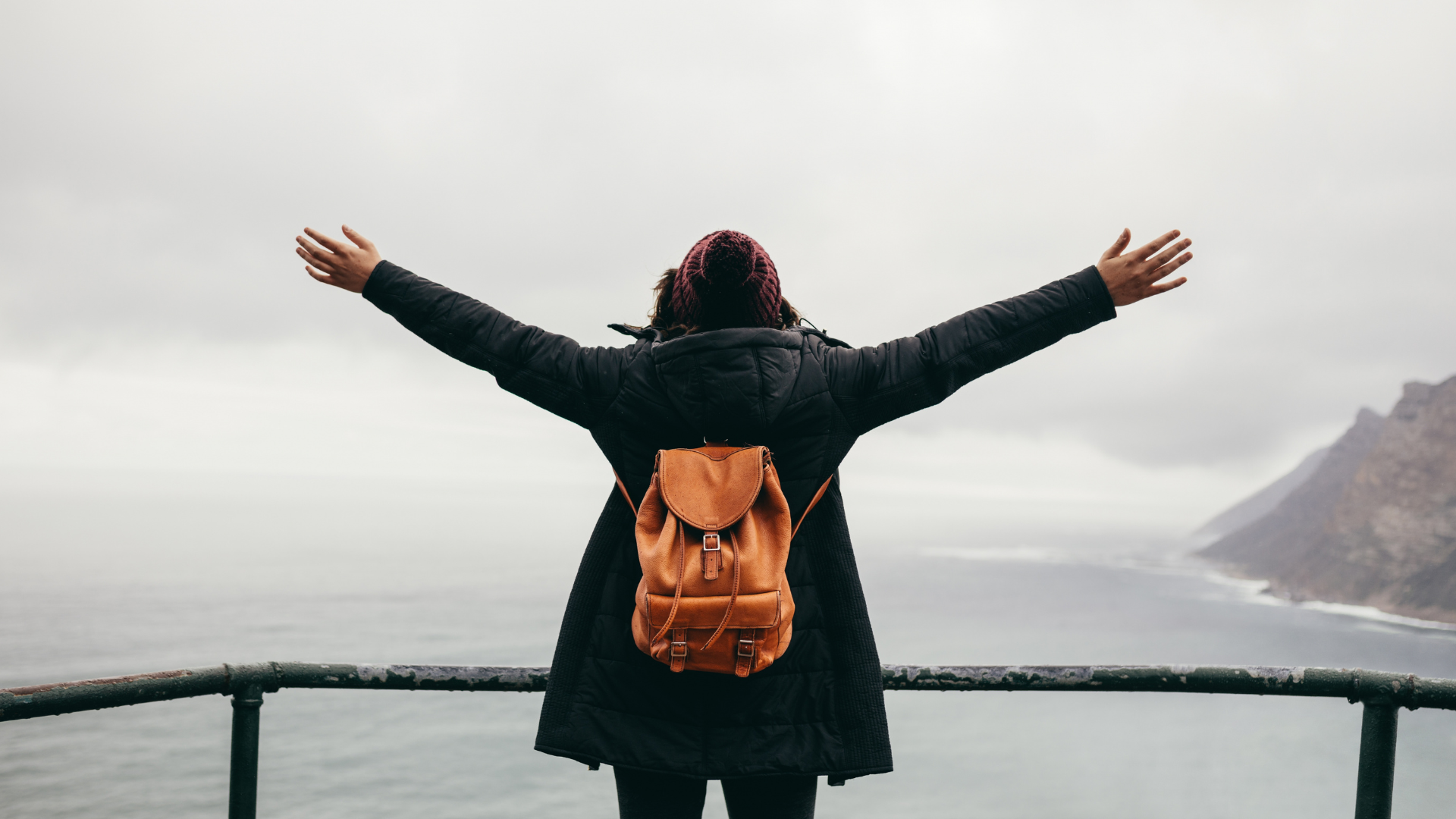 Traveler enjoying winter travel with arms wide open, wearing a black coat and backpack near a misty coastal mountain view
