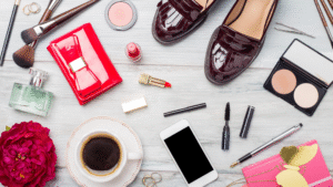 Flat lay of fashion accessories including shoes, red wallet, perfume, lipstick, makeup brushes, jewelry, and smartphone on a wooden background.