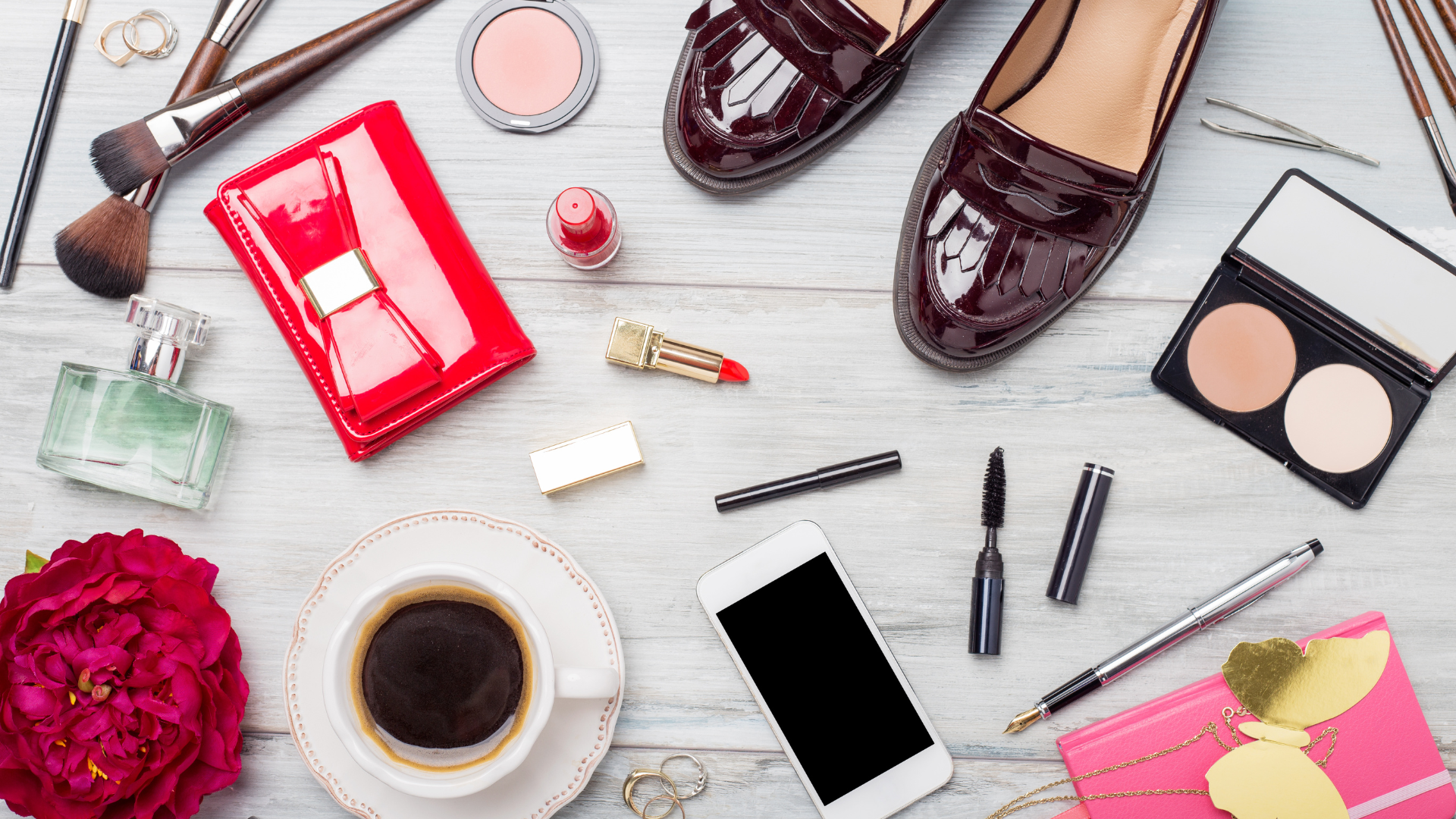 Flat lay of fashion accessories including shoes, red wallet, perfume, lipstick, makeup brushes, jewelry, and smartphone on a wooden background.