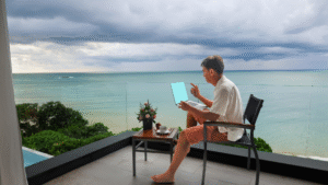 Digital nomad working remotely on a laptop with ocean view balcony, blending work and travel lifestyle.