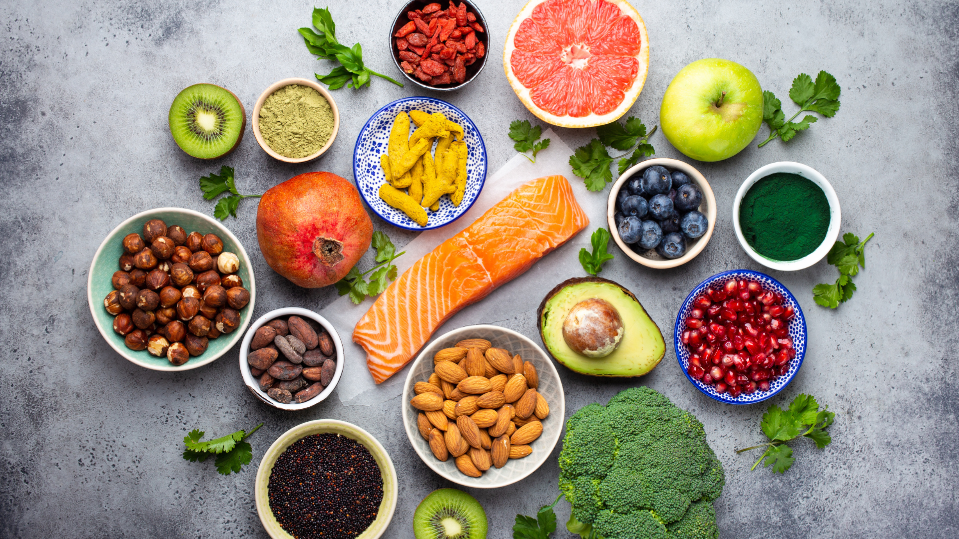 Assorted superfoods including salmon, avocado, nuts, berries, pomegranate, broccoli, and seeds arranged on a table for a healthy diet.
