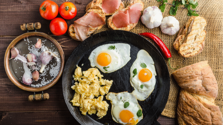 A variety of high-protein snacks including scrambled eggs, sunny-side-up eggs, garlic cloves, sliced ham on toast, tomatoes, and crusty bread arranged on a rustic wooden table.