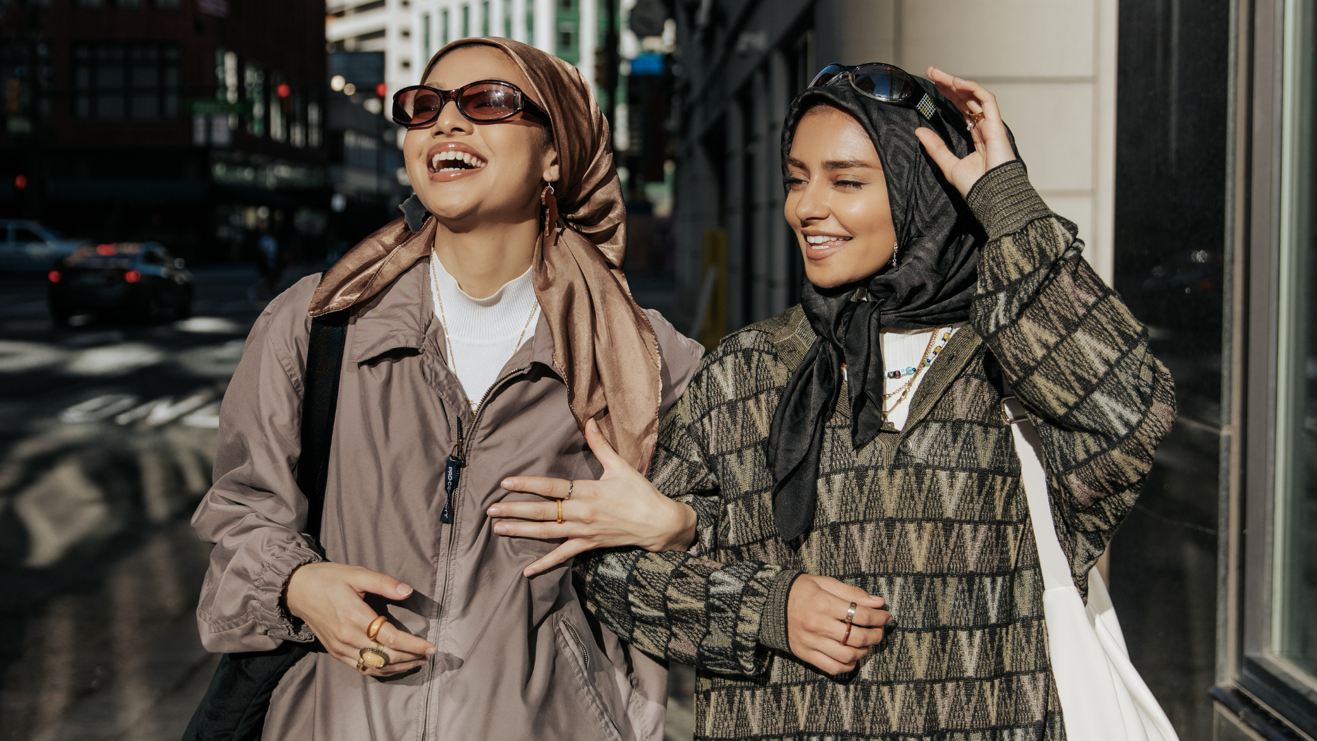Two stylish women in modern modest fashion wearing patterned jackets and sunglasses while walking in the city.