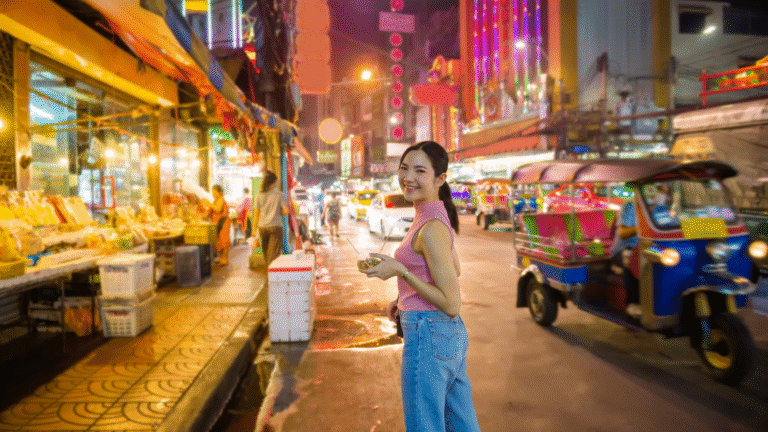 A smiling woman enjoying street foods at a vibrant, colorful night market street with food stalls and tuk-tuks in the background.