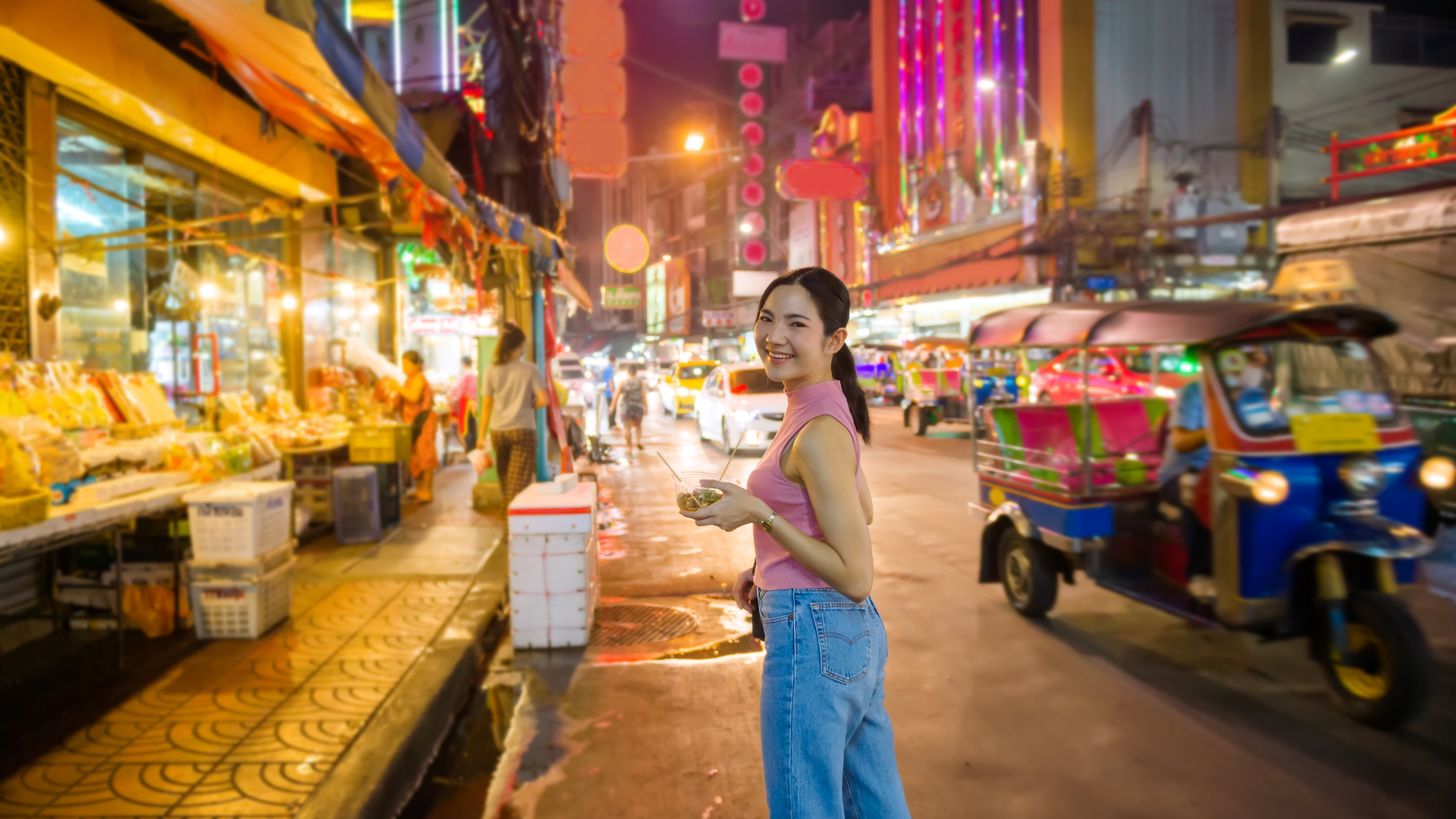 A smiling woman enjoying street foods at a vibrant, colorful night market street with food stalls and tuk-tuks in the background.