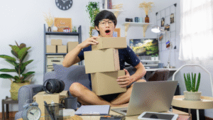 A young man sitting cross-legged in a home office, holding several cardboard boxes while working on his laptop, representing a dropshipping business setup.