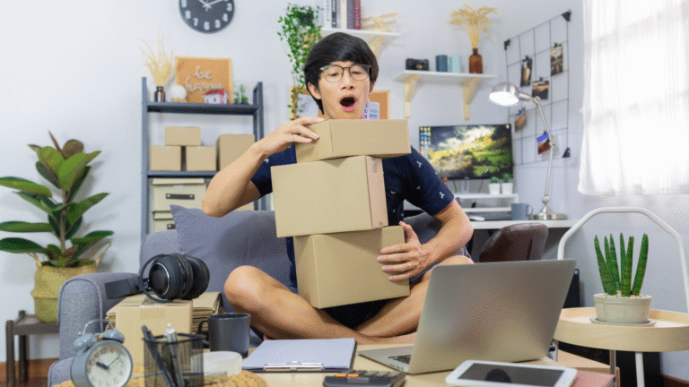 A young man sitting cross-legged in a home office, holding several cardboard boxes while working on his laptop, representing a dropshipping business setup.