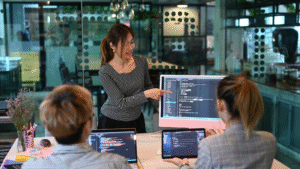 A woman standing and pointing at a computer monitor displaying code, explaining to two colleagues seated with laptops in a modern office setting, illustrating teamwork on finance tasks software development.