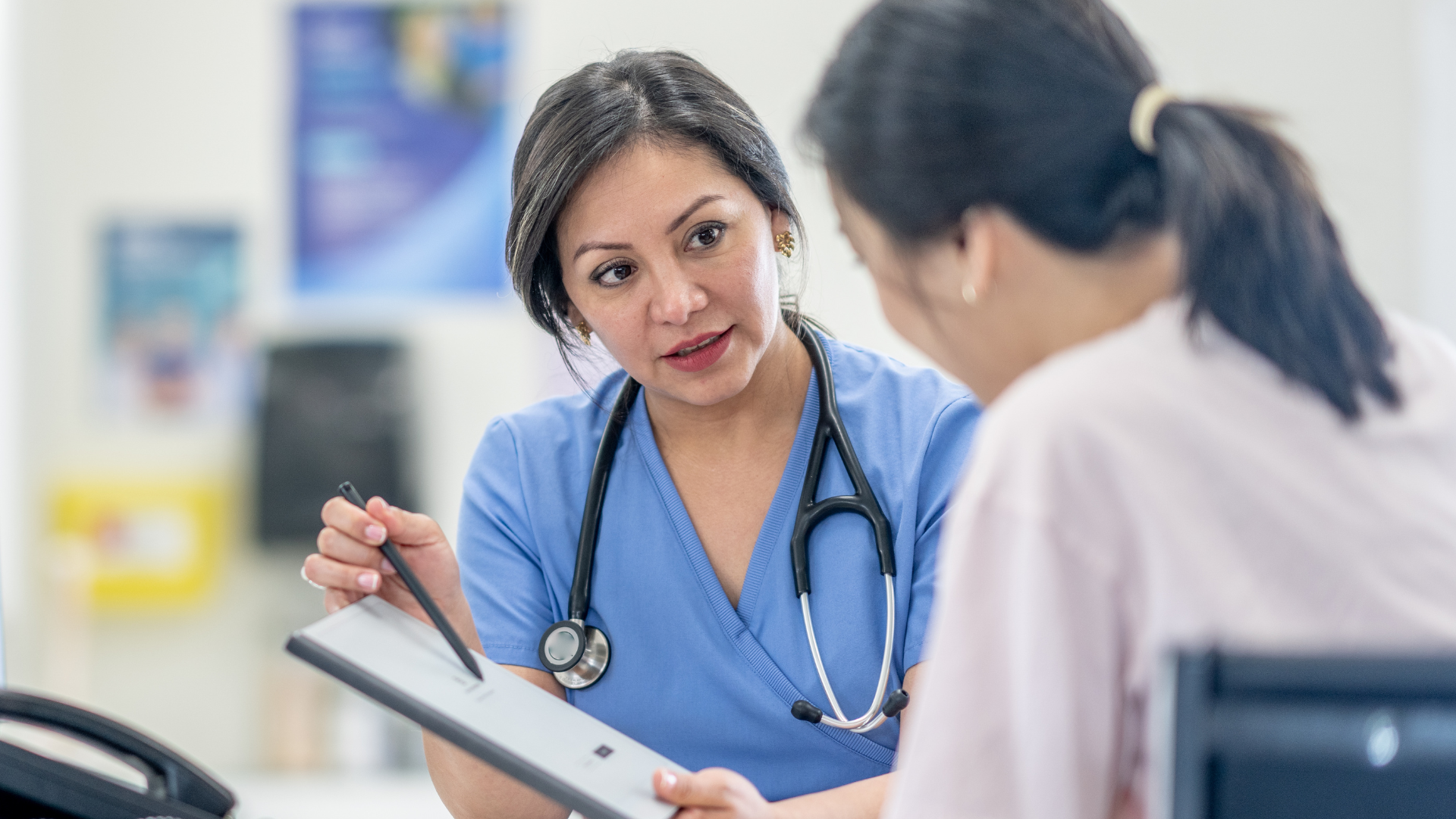 A female healthcare professional in blue scrubs with a stethoscope around her neck explains information on a clipboard to a patient, illustrating the importance of addressing common health myths through accurate medical advice