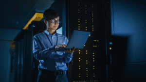 IT professional checking cybersecurity measures on a laptop in a server room with active data servers in the background