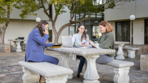 A diverse group of three professionals works together outdoors at a large stone picnic table. A man in a blue suit sits on the left, talking on his phone. In the middle, a woman in glasses and a gray jacket works on a laptop. On the right, another woman in a green sweater is looking at a notebook. The setting appears to be a corporate or university campus, with a building and trees in the background.