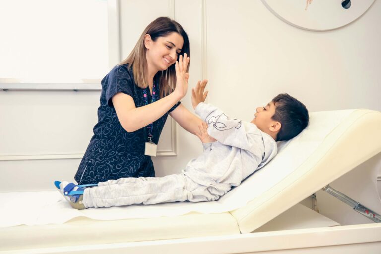 A cheerful pediatrician high-fives a young boy with a leg brace on an examination table, both smiling.