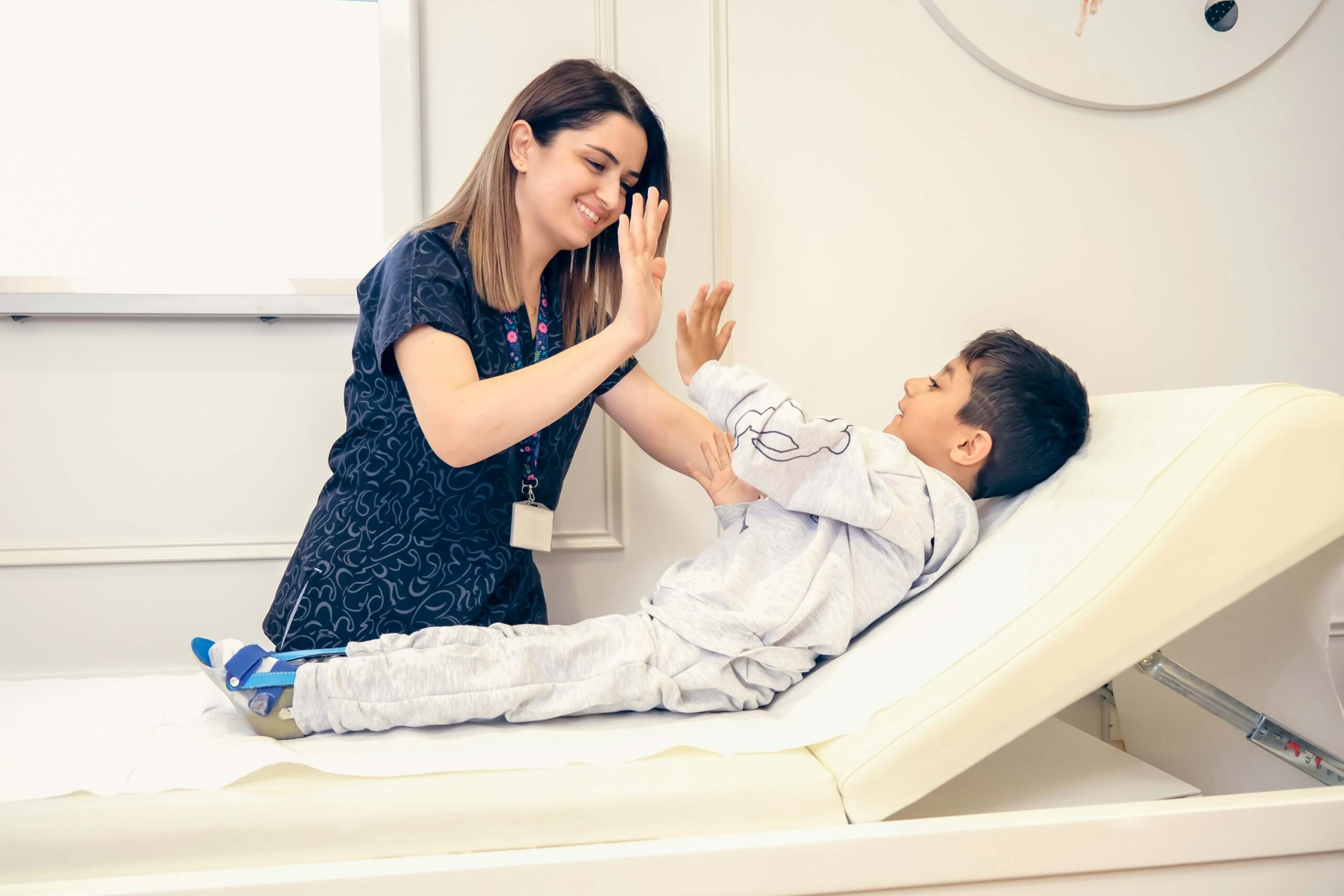 A cheerful pediatrician high-fives a young boy with a leg brace on an examination table, both smiling.