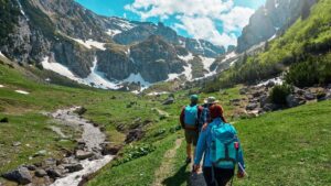 Adventure travel: Three hikers on a trail in a sunny green valley with towering, snow-dusted mountains in the background.