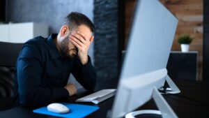 A stressed businessman covering his face while working on a computer, symbolizing frustration and anxiety caused by data breaches and cybersecurity issues in the workplace.