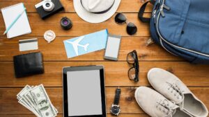 Flat lay of essential travel gadgets including a camera, smartphone, tablet, compass, sunglasses, wallet, and backpack arranged on a wooden surface.