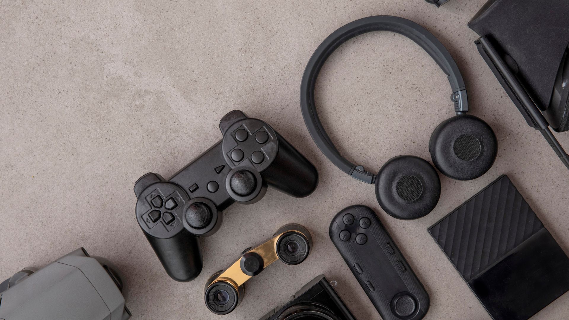 Flat lay of modern tech gadgets including a gaming controller, wireless headphones, remote device, portable camera gear, and accessories arranged on a neutral surface.