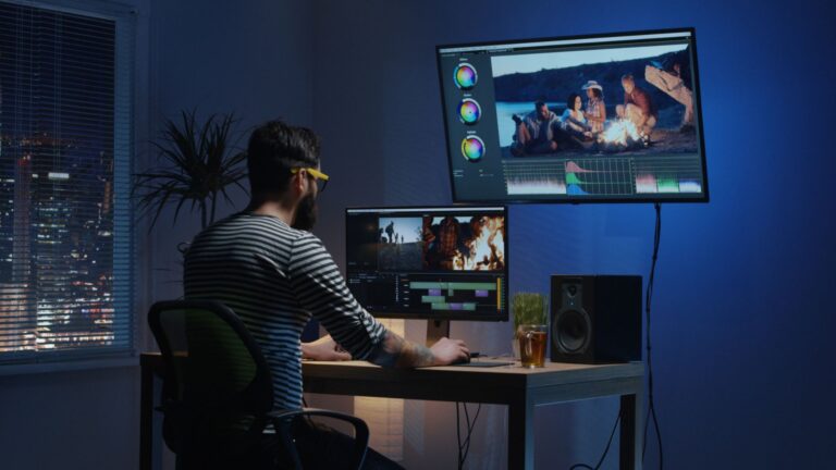 A man working on a dual-monitor setup at night, editing footage using video editing apps, with color grading tools and timeline clips visible on the screens.