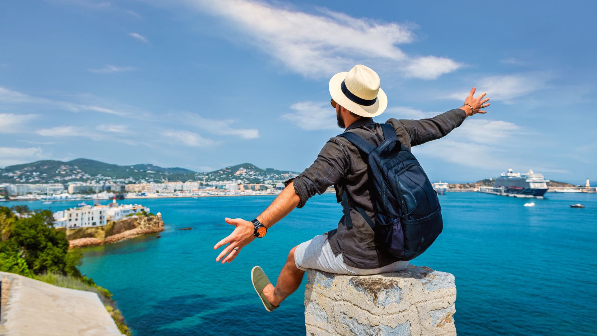 Traveler sitting on a seaside cliff with open arms overlooking blue water and a coastal city, representing travel trends and modern travel experiences.
