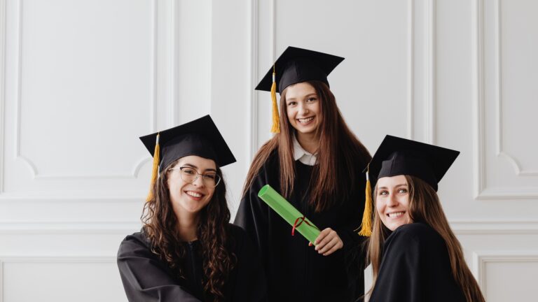 Three smiling female graduates wearing black graduation gowns and mortarboards pose indoors, one holding a green diploma scroll tied with a ribbon—representing Scholarship Hunting: Tips to Secure Funding for Higher Education through academic achievement, financial aid opportunities, and successful graduation outcomes.
