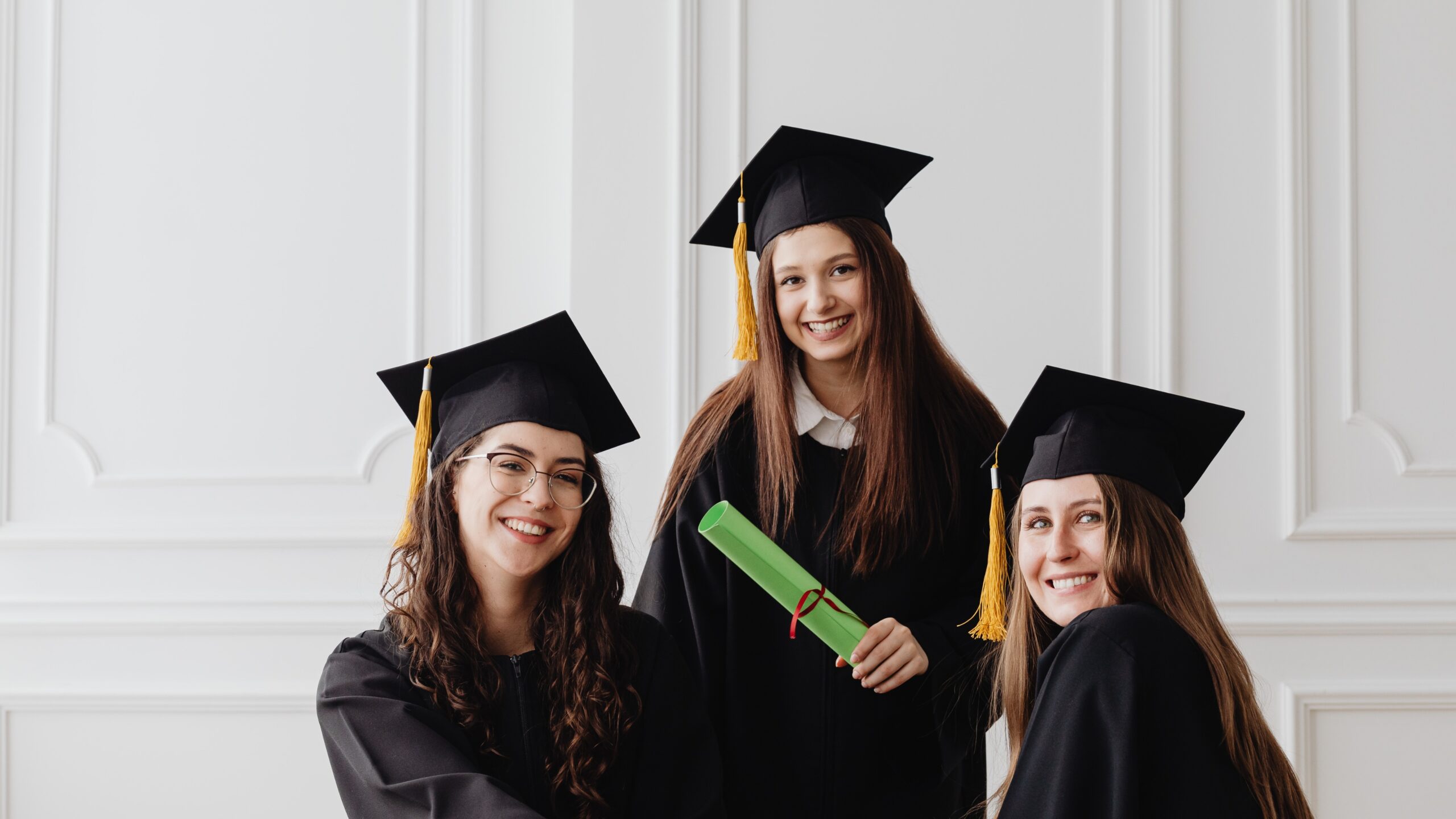 Three smiling female graduates wearing black graduation gowns and mortarboards pose indoors, one holding a green diploma scroll tied with a ribbon—representing Scholarship Hunting: Tips to Secure Funding for Higher Education through academic achievement, financial aid opportunities, and successful graduation outcomes.