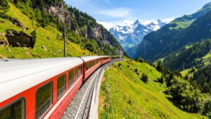 A red panoramic train curves along a mountainside through lush green valleys with snow-capped Alpine peaks in the distance—capturing Train Journeys Around the World: Scenic Routes from the Rockies to the Alps and the beauty of long-distance rail travel through dramatic landscapes.
