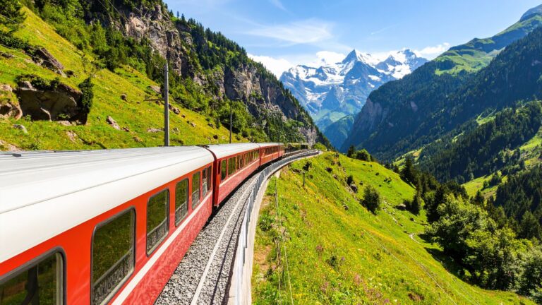 A red panoramic train curves along a mountainside through lush green valleys with snow-capped Alpine peaks in the distance—capturing Train Journeys Around the World: Scenic Routes from the Rockies to the Alps and the beauty of long-distance rail travel through dramatic landscapes.
