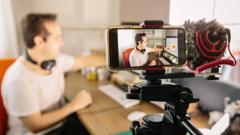 Content creator filming a YouTube video using a smartphone setup with a microphone in a home studio.