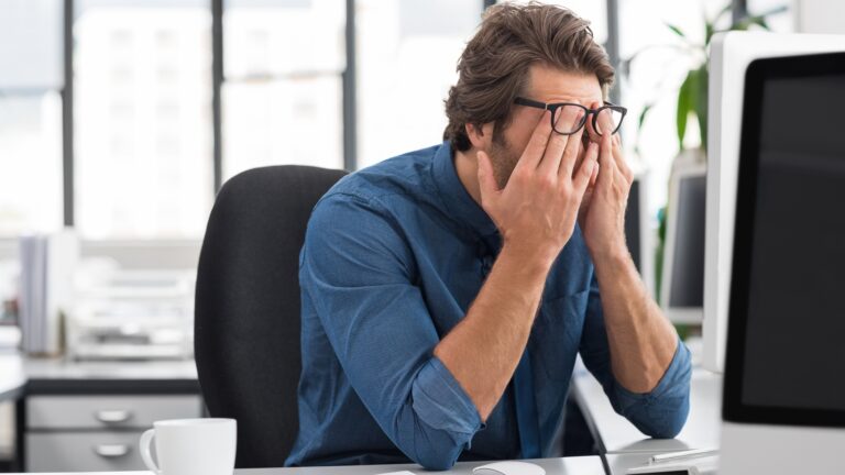 Man experiencing fatigue and tension at his desk, rubbing his eyes while working on a computer, illustrating health problems caused by long-term stress such as burnout, headaches, and mental exhaustion.