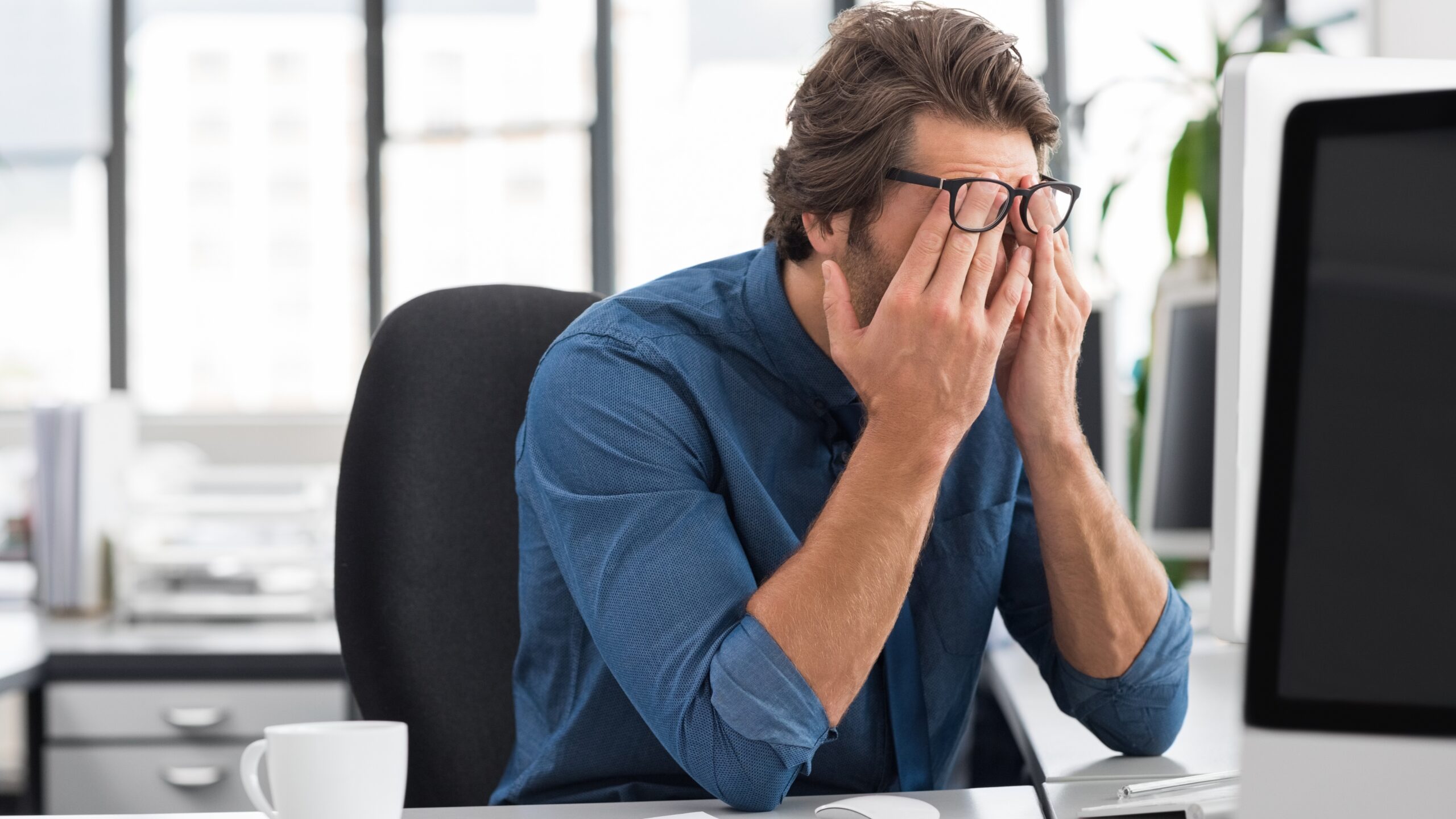 Man experiencing fatigue and tension at his desk, rubbing his eyes while working on a computer, illustrating health problems caused by long-term stress such as burnout, headaches, and mental exhaustion.