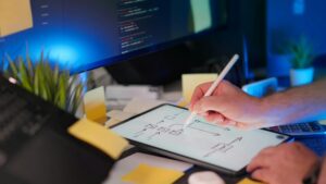 The Role of Open-Source Software in Modern App Development shown through a developer sketching app architecture on a tablet with code displayed on a desktop monitor.