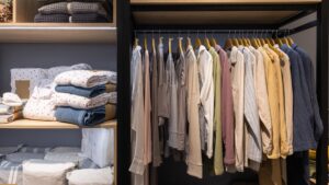 Modern wardrobe with neatly organized clothes, folded linens on shelves, and neutral-toned shirts and jackets hanging on wooden hangers in a minimalist closet setup.