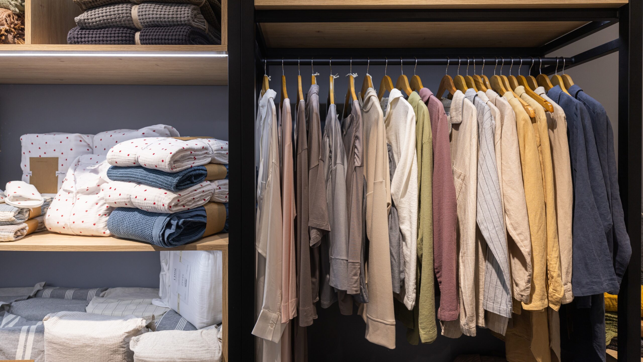 Modern wardrobe with neatly organized clothes, folded linens on shelves, and neutral-toned shirts and jackets hanging on wooden hangers in a minimalist closet setup.