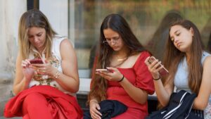Three women sitting outdoors using their smartphones, illustrating Top Smartphone Brands Compared: iPhone vs Android in 2026 Performance.